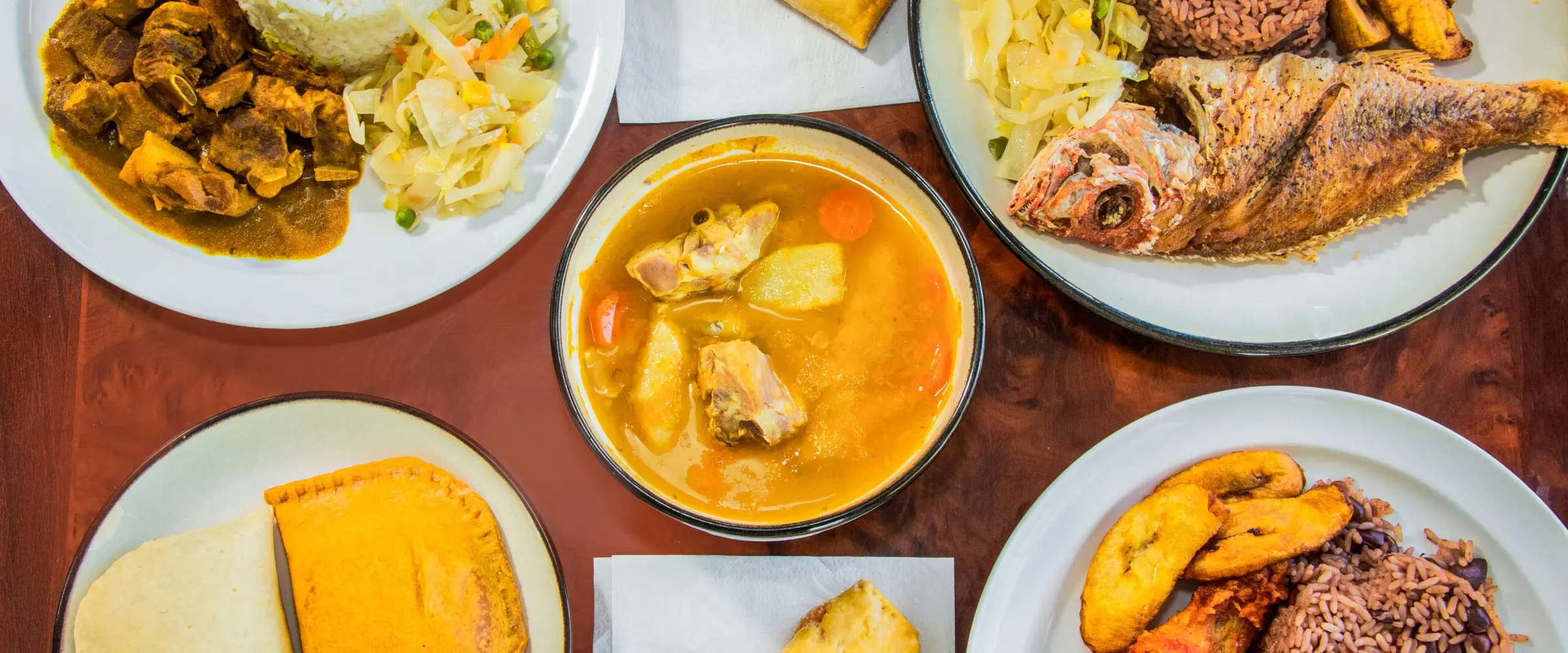 A spread of Nice and Knuff dishes: curry goat, fried snapper, soup, beef patties with coco bread, and plantains with rice and peas